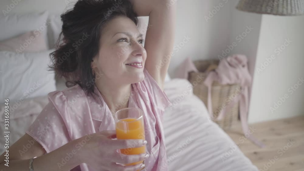 Face of beautiful brunette Caucasian woman adjusting hair and smiling. Happy lady with orange juice in hand enjoying morning on vacations indoors. Portrait of woman in pink pajamas. Lifestyle. Face of beautiful brunette Caucasian woman adjusting hair and smiling. Happy lady with orange juice in hand enjoying morning on vacations indoors. Portrait of woman in pink pajamas. Lifestyle.