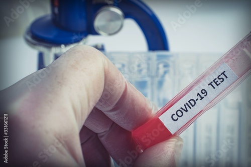 A hand in a protective glove holds a test tube labeled COVID-19 with red liquid against the background of a microscope and a number of clean medical tubes. The problem of the spread of coronavirus inf