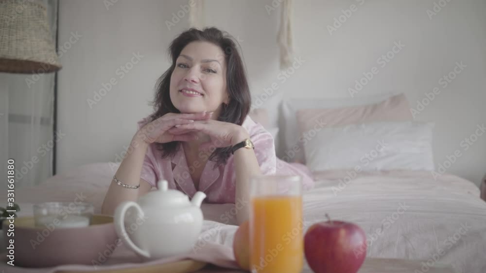 Portrait of positive Caucasian brunette woman posing in the morning in bedroom. Charming young lady lying on bed and smiling. Leisure, resting, weekends, lifestyle. Portrait of positive Caucasian brunette woman posing in the morning in bedroom. Charming young lady lying on bed and smiling. Leisure, resting, weekends, lifestyle.