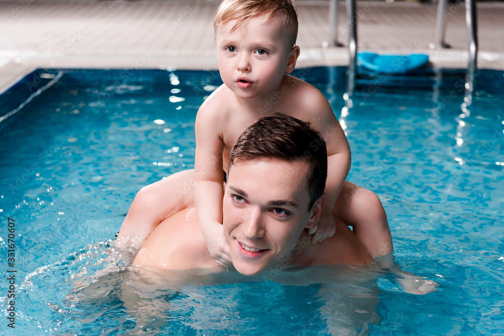 handsome swim coach holding cute toddler kid on shoulders in swimming pool
