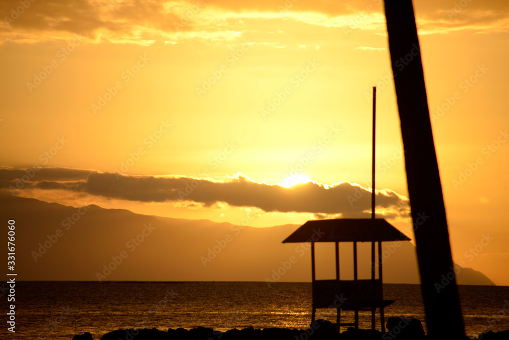 Atardecer anaranjado en la costa con una caseta de socorristas y el tronco de una palmera