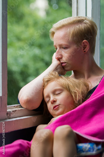 Mother and daughter are sitting by the open window, the mother is holding her sleeping daughter in a blanket on herself, the mother is looking out the window with alarm.