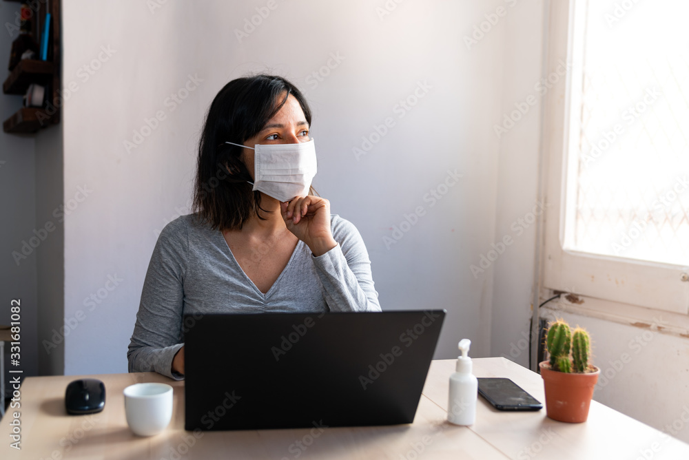 young woman wearing protective face mask and with hand sanitizer in home office looking worried and stressed out of the window due to corona virus young woman wearing protective face mask and with hand sanitizer in home office looking worried and stressed out of the window due to corona virus