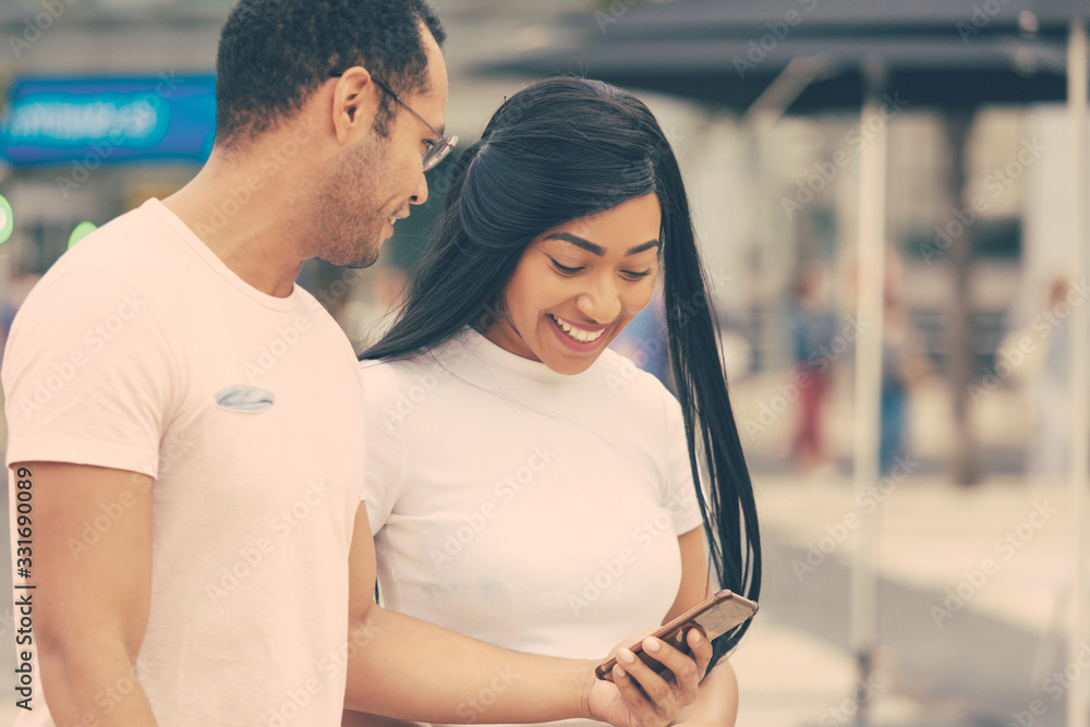 Cheerful young friends looking at smartphone during stroll. Front view of happy couple walking on street and talking. Communication and technology concept Cheerful young friends looking at smartphone during stroll. Front view of happy couple walking on street and talking. Communication and technology concept