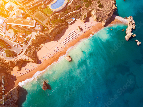 View of stunning turquoise beach, hotel, houses, a pool and tennis field on top of golden color rocks, Algarve, Portugal. 