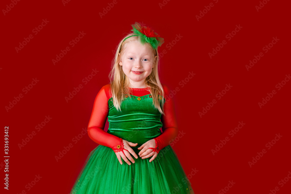 Little blonde girl in a christmas and new year green costume having fun and sending kisses on red background. Space for text. Dreams come true Merry Christmas Little blonde girl in a christmas and new year green costume having fun and sending kisses on red background. Space for text. Dreams come true Merry Christmas
