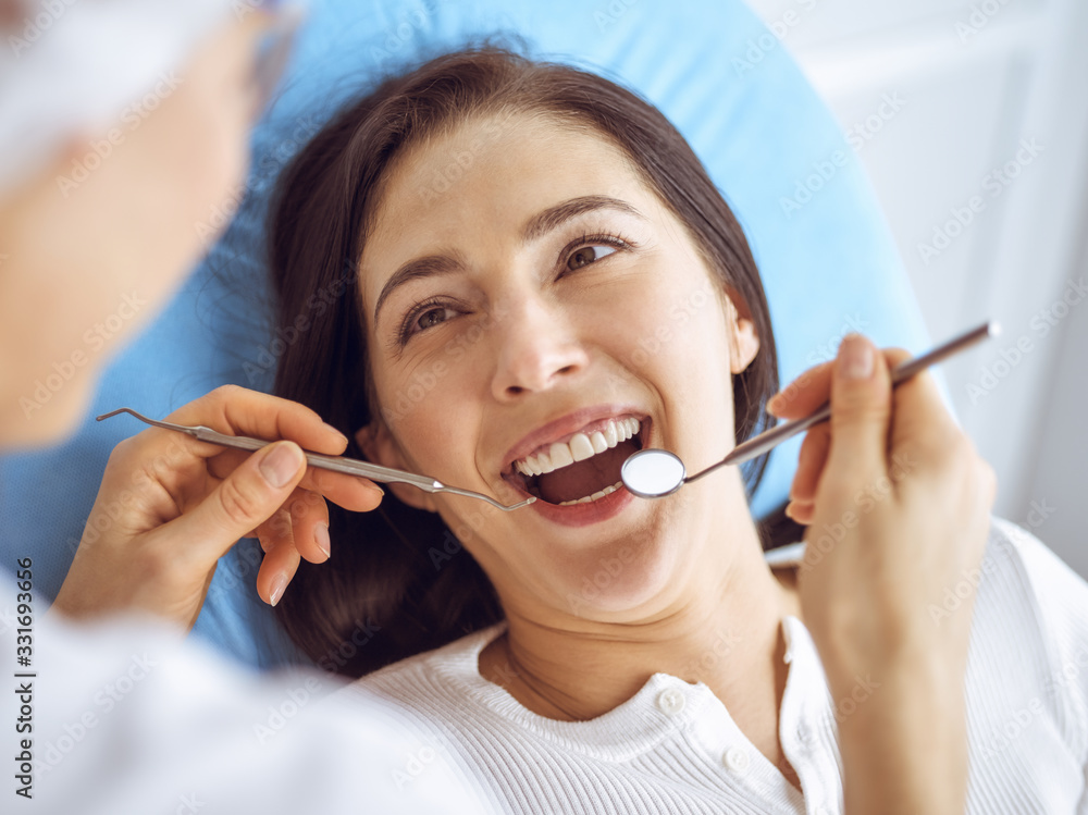 Smiling brunette woman being examined by dentist at dental clinic. Hands of a doctor holding dental instruments near patient's mouth. Healthy teeth and medicine concept Smiling brunette woman being examined by dentist at dental clinic. Hands of a doctor holding dental instruments near patient's mouth. Healthy teeth and medicine concept