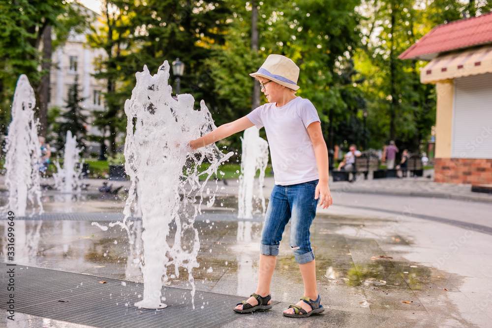 Obraz premium Little caucasian boy in hat playing and having fun with water in fountain in the sunny summer park