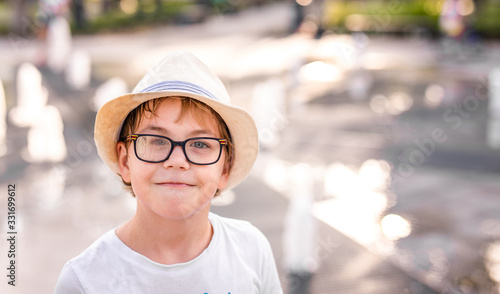 Little caucasian boy in hat and big glasses playing and having fun with water in fountain in the sunny summer park.