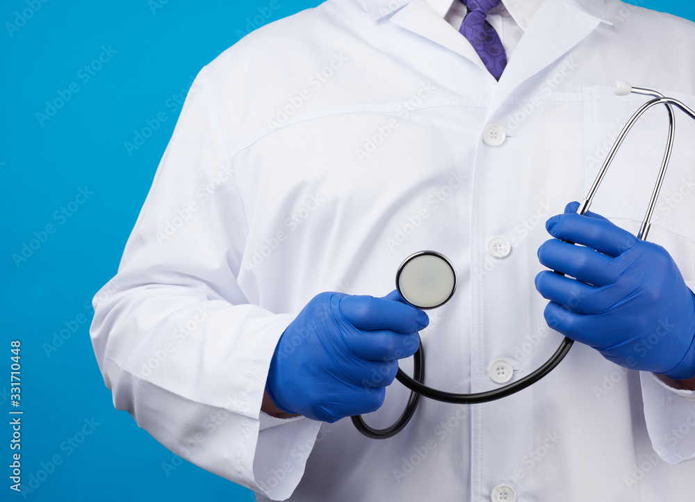 adult male doctor in a white medical coat stands and holds a black stethoscope on a blue background adult male doctor in a white medical coat stands and holds a black stethoscope on a blue background