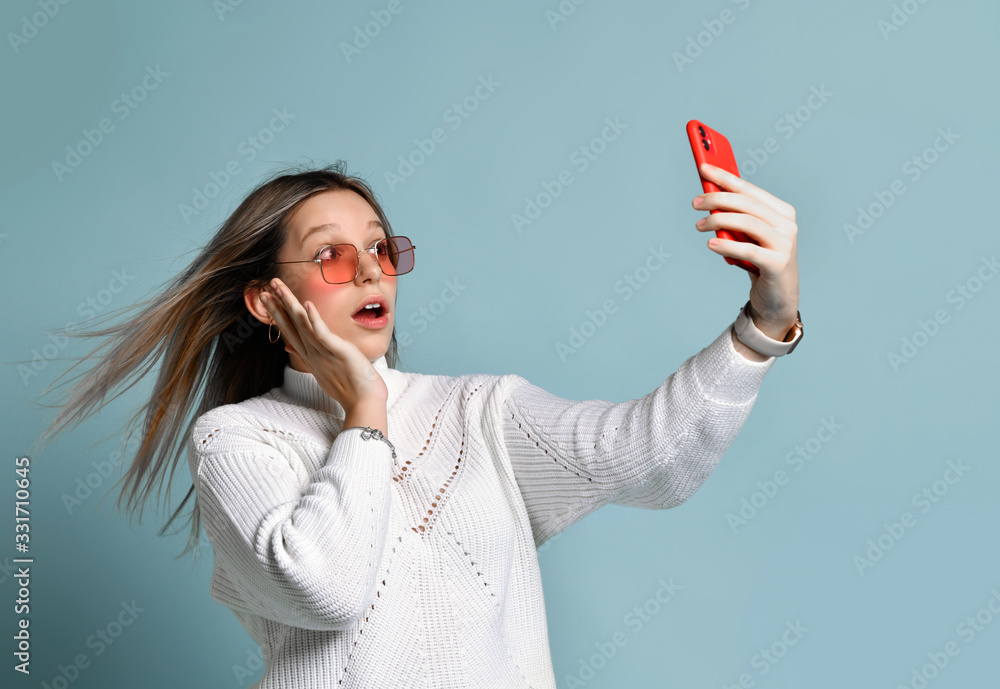 Teenager in orange sunglasses, watch, bracelet and sweater. She taking selfie, surprised, posing on blue background. Close up Teenager in orange sunglasses, watch, bracelet and sweater. She taking selfie, surprised, posing on blue background. Close up