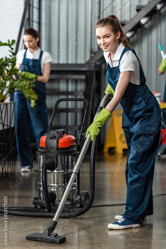 selective focus of smiling cleaner vacuuming floor near colleague cleaning plants