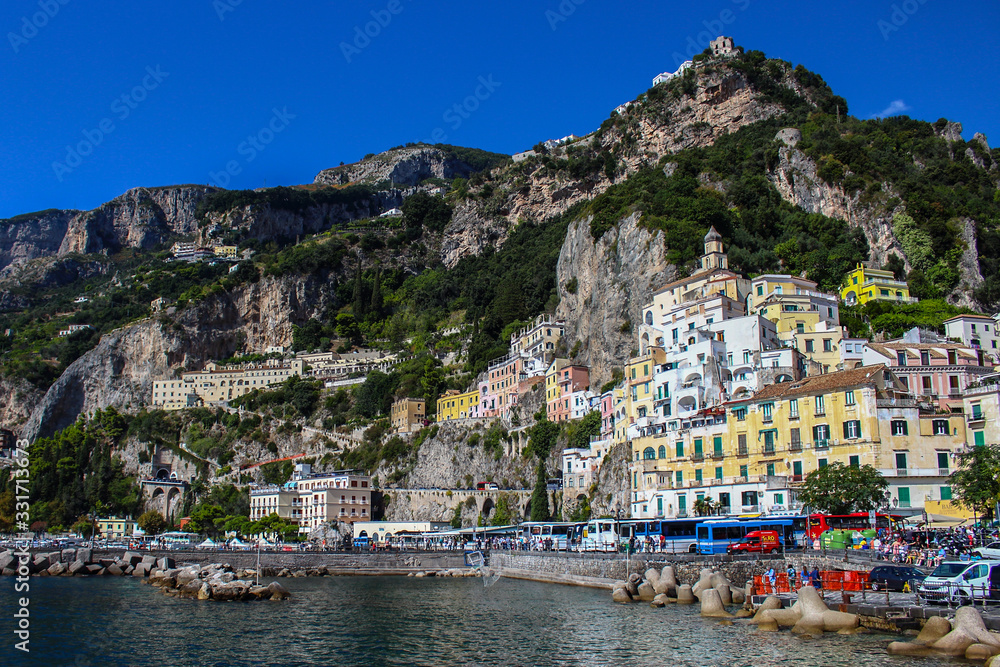 View of the city of Amalfi from the jetty with parked buses, the sea and the colorful houses on the slopes of the Amalfi coast in the province of Salerno, Campania, Italy.