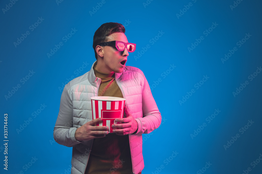 Shocked in 3D eyewear and with popcorn. Caucasian man's portrait isolated on blue studio background in pink neon light. Beautiful male model. Concept of human emotions, facial expression, sales, ad. Shocked in 3D eyewear and with popcorn. Caucasian man's portrait isolated on blue studio background in pink neon light. Beautiful male model. Concept of human emotions, facial expression, sales, ad.