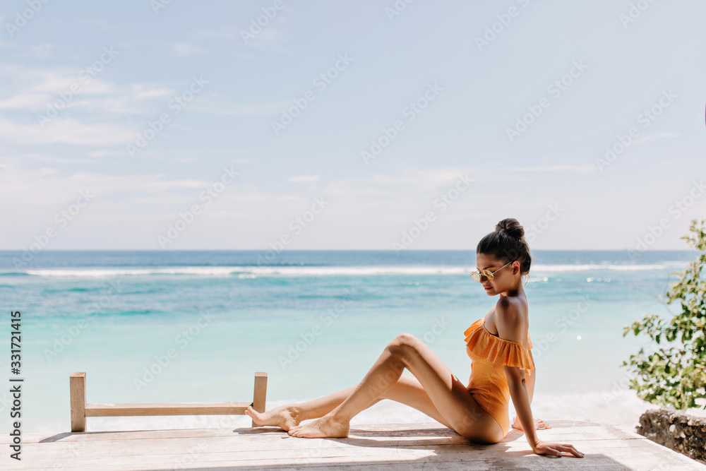 Romantic tanned girl in orange attire sitting on ocean background. Enchanting white female model in sunglasses posing on the ground on sea coast. Romantic tanned girl in orange attire sitting on ocean background. Enchanting white female model in sunglasses posing on the ground on sea coast.