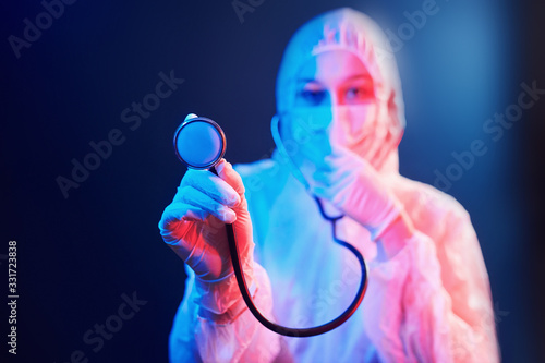 Nurse in mask and white uniform and with stethoscope standing in neon lighted room