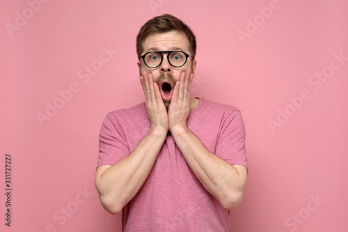 Cute man in funny glasses is surprised, he holds face with hands, in shock opened mouth and looks at the camera.