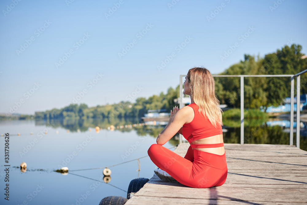 Young blond woman, wearing red fitness outfit sitting on wooden pier dock in summer morning. Healthy active life concept. Portrait of girl resting after training exercise outside in the city. Young blond woman, wearing red fitness outfit sitting on wooden pier dock in summer morning. Healthy active life concept. Portrait of girl resting after training exercise outside in the city.