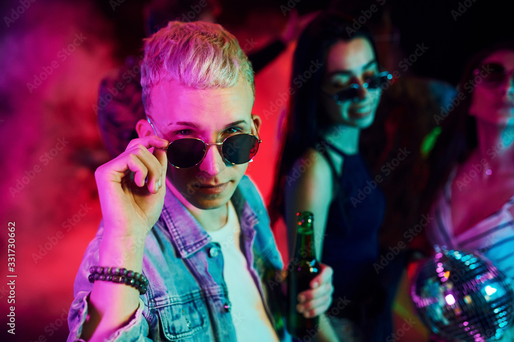Hipster guy in sunglasses and with bottle of alcohol posing for camera in front of young people that having fun in night club with colorful laser lights