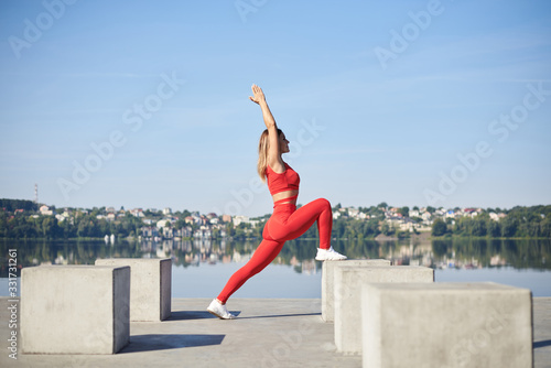 Young blond woman, wearing red fitness outfit standing on concrete platform by city lake. Healthy active life concept. Portrait of girl training, doing sports stretching exercise outside in summer.