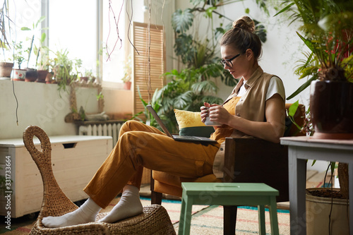 Young woman in eyeglasses sitting on armchair drinking coffee and watching something on laptop in the room with plants