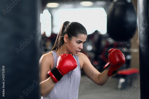 Athletic young brunette woman in sportswear and red boxing gloves trains bumps on a punching bag in a fitness gym.