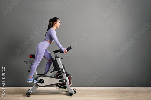 A beautiful athletic young brunette woman in sportswear trains on a sycle in the gym against the gray wall.