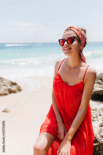 Laughing tanned white woman sitting at beach. Outdoor portrait of inspired caucasian female model in red dress spending leisure time near sea.