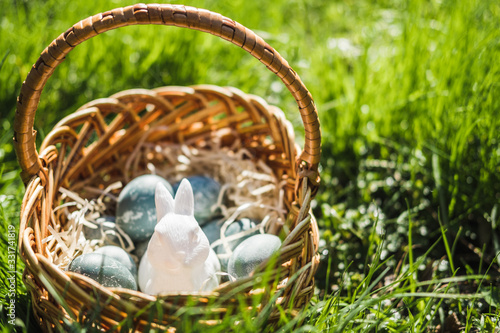 Painted blue textured easter eggs and a white plastic rabbit in a wicker brown hand made basket on green fresh grass. The concept of the spring holiday and egg hunting. photo