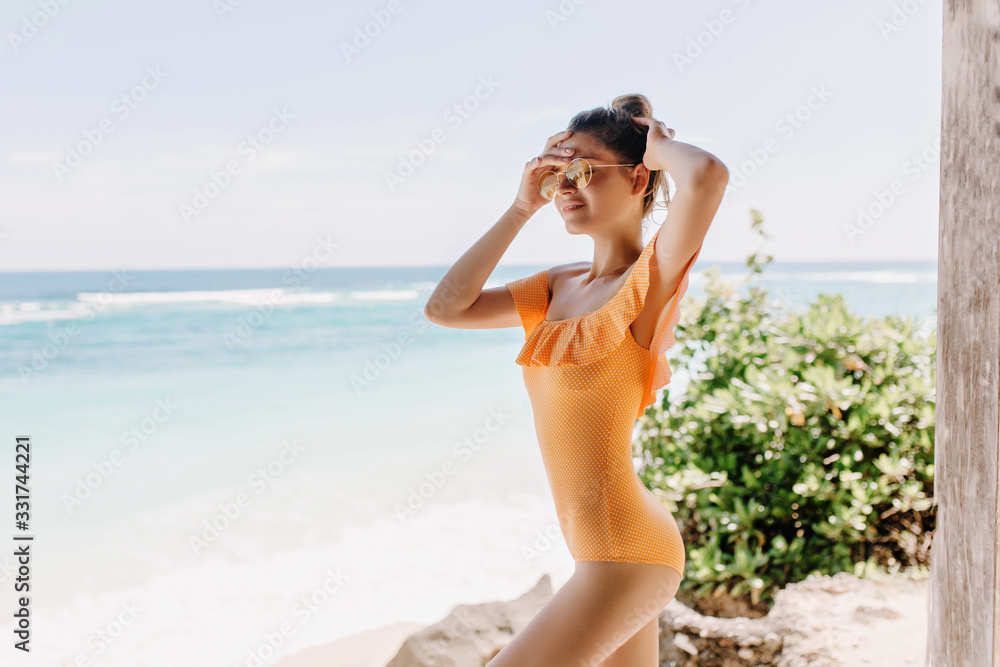 Graceful white female model in yellow clothes posing on ocean background. Slim caucasian girl in orange swimsuit chilling near sea with eyes closed. Graceful white female model in yellow clothes posing on ocean background. Slim caucasian girl in orange swimsuit chilling near sea with eyes closed.
