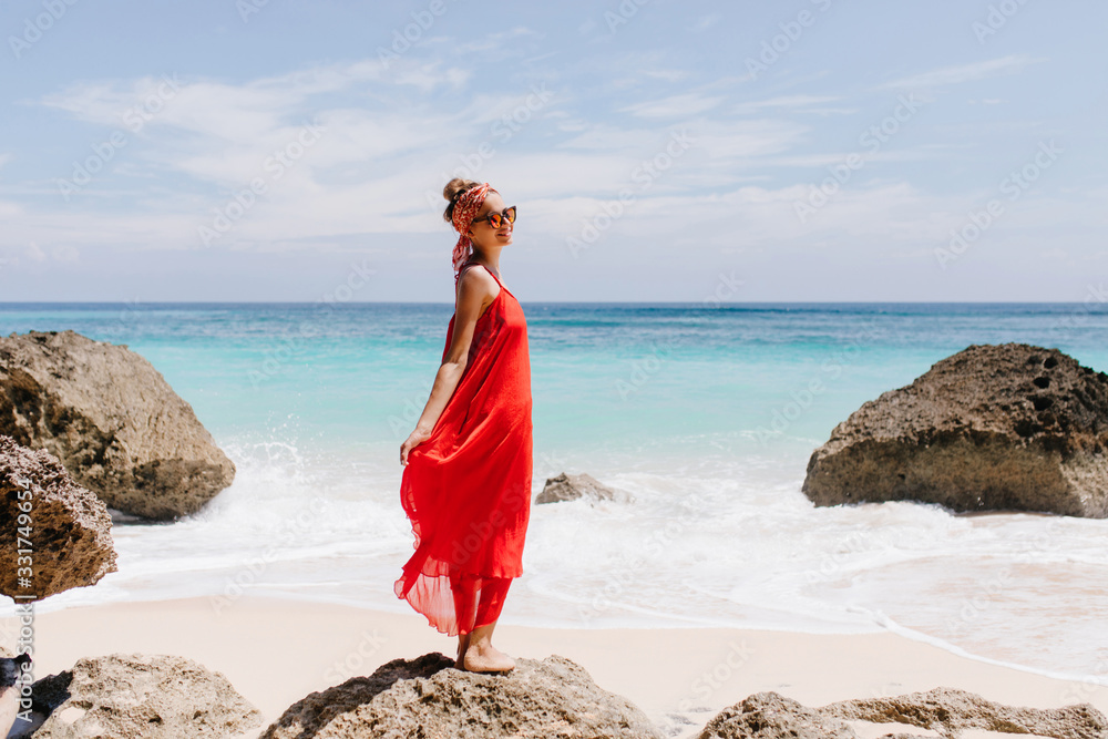Ecstatic girl with pretty smile standing on big stone with ocean on background. Outdoor full-length photo of cheerful female tourist chilling at wild beach. Ecstatic girl with pretty smile standing on big stone with ocean on background. Outdoor full-length photo of cheerful female tourist chilling at wild beach.