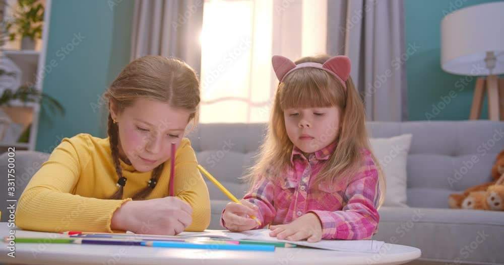 Caucasian little pretty and cute sisters sitting in cozy nice room at desk and coloring picture with colorful pencils. Joyful girls having fun and play indoors. Kids spending time alone at home. Caucasian little pretty and cute sisters sitting in cozy nice room at desk and coloring picture with colorful pencils. Joyful girls having fun and play indoors. Kids spending time alone at home.