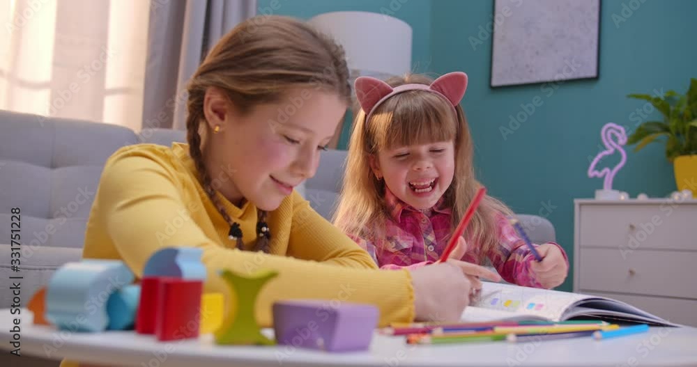 Cheerful small Caucasian sisters sitting at home alone and playing nice together. Cute kids coloring picture with colorful pencils in cozy room. Happy children play and talking indoor. Cheerful small Caucasian sisters sitting at home alone and playing nice together. Cute kids coloring picture with colorful pencils in cozy room. Happy children play and talking indoor.