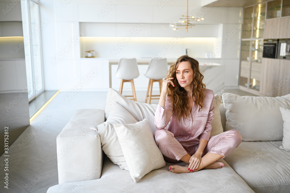 Beautiful young woman with curly hair in pink pajamas sitting at white couch sofa in the morning. Scandinavian style living room & kitchen interior. Enjoying time at home. Furniture store concept Beautiful young woman with curly hair in pink pajamas sitting at white couch sofa in the morning. Scandinavian style living room & kitchen interior. Enjoying time at home. Furniture store concept