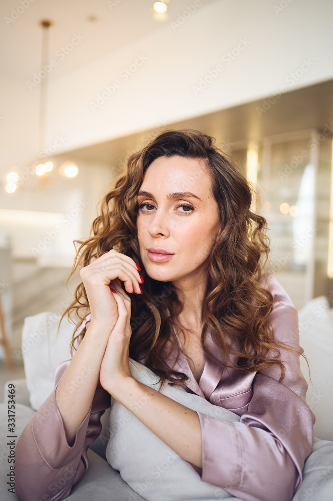 Beautiful young woman with curly hair in pink pajamas sitting at white couch or sofa in the morning. Scandinavian style living room & kitchen interior. Enjoying time at home. Furniture store concept Beautiful young woman with curly hair in pink pajamas sitting at white couch or sofa in the morning. Scandinavian style living room & kitchen interior. Enjoying time at home. Furniture store concept