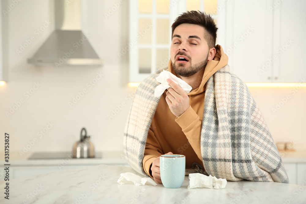 Sick young man with cup of hot drink and tissues in kitchen. Influenza virus Sick young man with cup of hot drink and tissues in kitchen. Influenza virus