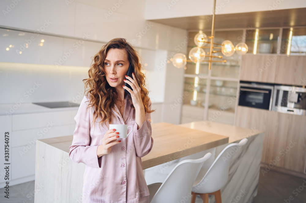 Beautiful young woman with curly hair drinking coffee at home in lazy weekend morning in cozy pink pajamas. Lady speaking phone in scandinavian style white kitchen interior. Distance work from home Beautiful young woman with curly hair drinking coffee at home in lazy weekend morning in cozy pink pajamas. Lady speaking phone in scandinavian style white kitchen interior. Distance work from home