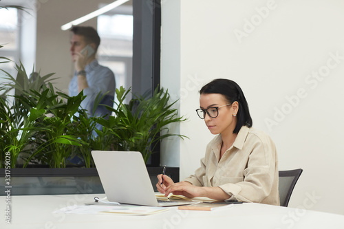 Serious manager in eyeglasses concentrating on her online work she working on laptop computer at office