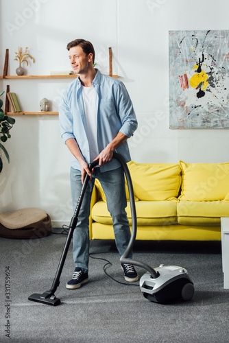 Smiling man looking away while cleaning carpet with vacuum cleaner at home