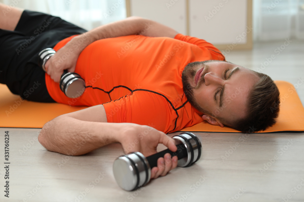 Lazy young man with sport equipment on yoga mat at home - obrazy, fototapety, plakaty 