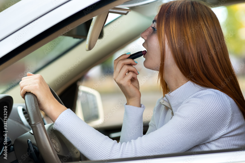 Closeup of a young redhead woman driver correcting her makeup with dark red lipstick looking in car rearview mirror behind steering wheel of a vehicle.