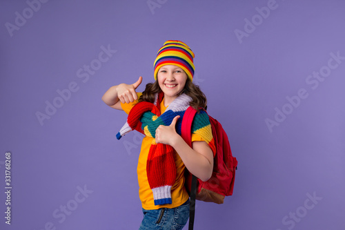 Schoolgirl girl smiles in yellow T-shirt, knitted sweater and multi-colored hat with a backpack on her shoulder on a purple background