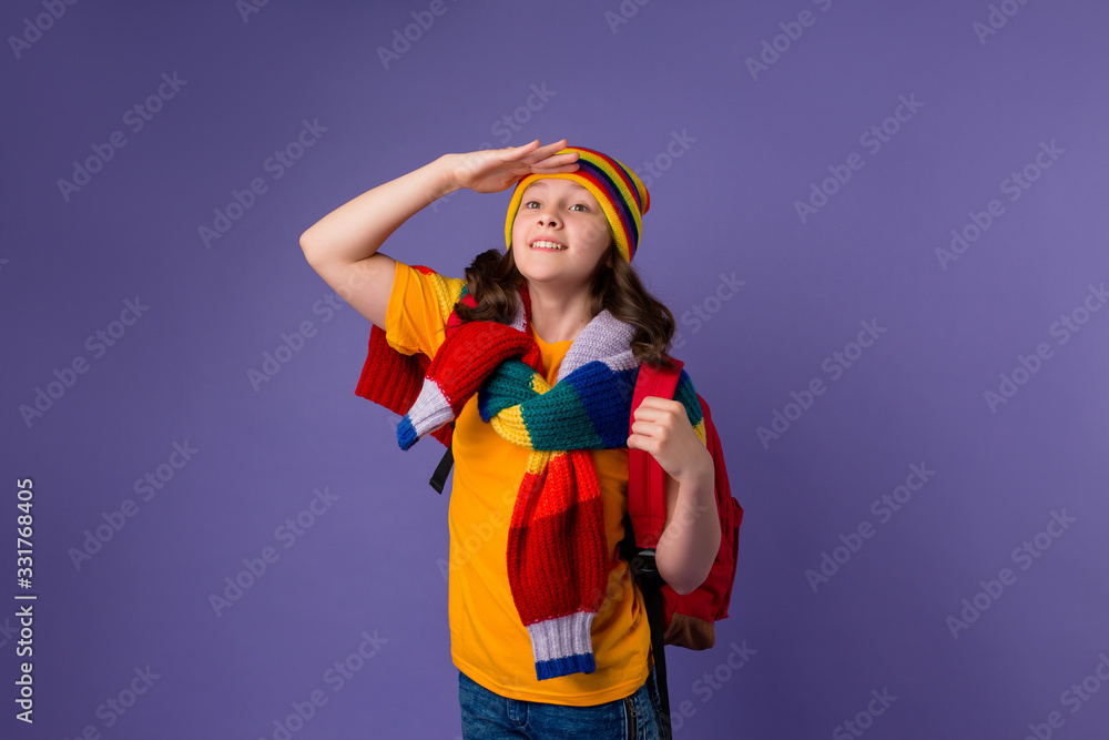 Schoolgirl girl smiles in yellow T-shirt, knitted sweater and multi-colored hat with a backpack on her shoulder on a purple background Schoolgirl girl smiles in yellow T-shirt, knitted sweater and multi-colored hat with a backpack on her shoulder on a purple background