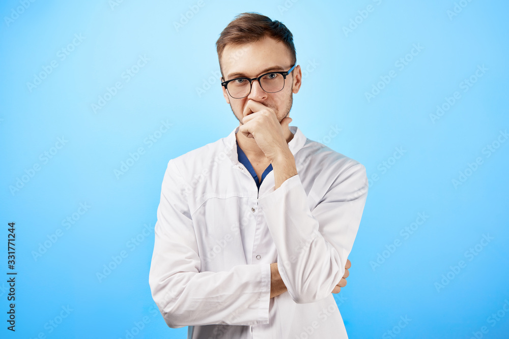 Portrait of intern doctor in white medical coat and glasses with a serious face expression isolated on blue wall background Portrait of intern doctor in white medical coat and glasses with a serious face expression isolated on blue wall background