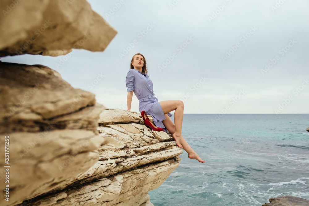 Sexy caucasian woman in striped dress sits at the edge of cliff with red shoes. Female tourist at rock by the sea in cloudy weather.