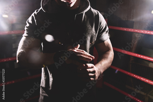 Closeup portrait professional boxer in a hood wraps the palms of his hands, puts on gloves in the ring. Dark colors, no face
