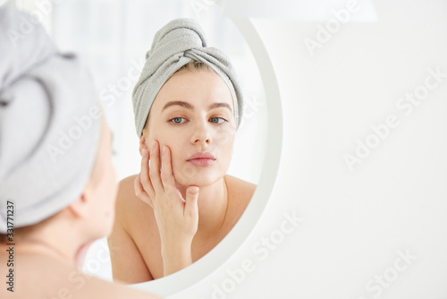 Portrait of young girl with towel on head in white bathroom looks and touches her face in the mirror and enjoys youth and hydration. Natural beauty, home care for problem skin