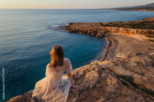 A beautiful young woman in pink dress sits at the edge of cliff and feeling sea breeze at her relaxing face. Sunset by the sea. © flagmen