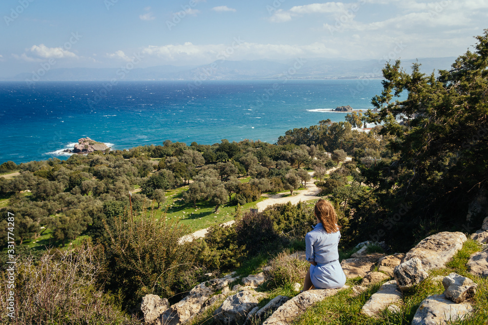 Attractive young girl in a striped dress sits on the rock and looking at panoramic view to the sea, trees and mountains. Sunny but windy weather