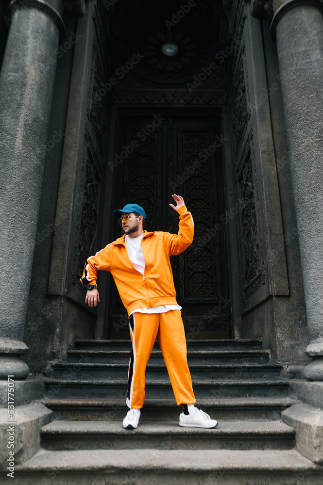 Stylish street dancer in bright casual clothes shows a hip hop performance against a backdrop of dark fringe. Man in a hat, glasses and orange tracksuit dancing hip hop on the street. Vertical.